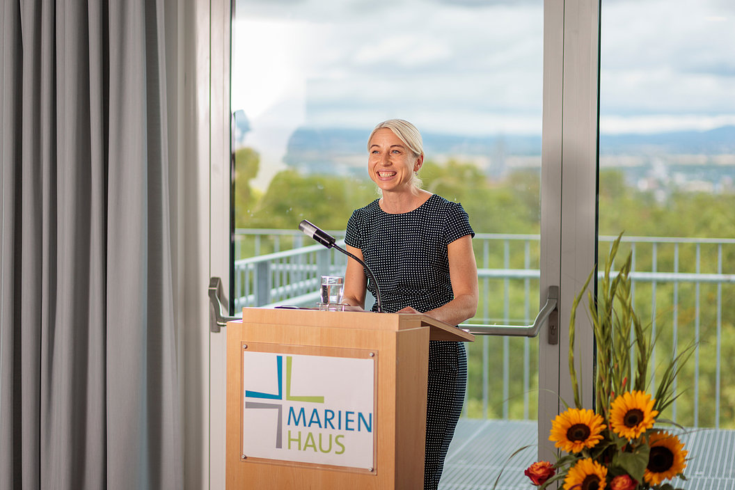 Frau mit blondem Haar spricht lächelnd an einem Rednerpult mit Mikrofon und Glas Wasser, vor Fenster mit Blick auf grüne Landschaft, Schild 'Marienhaus' am Pult